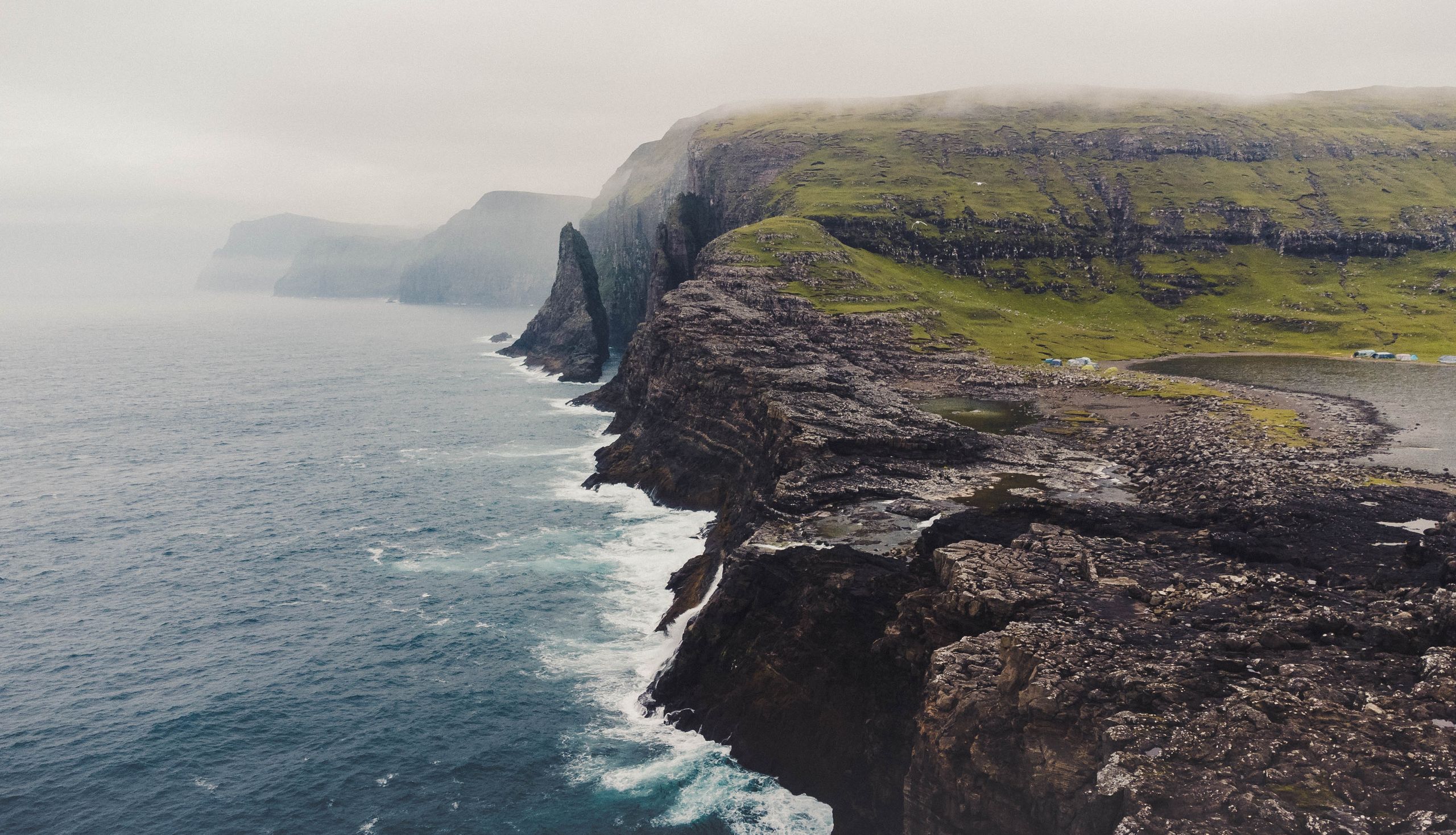 Aerial view of misty cliffs meeting the sea in Vága, Faroe Islands, capturing nature's rugged beauty.