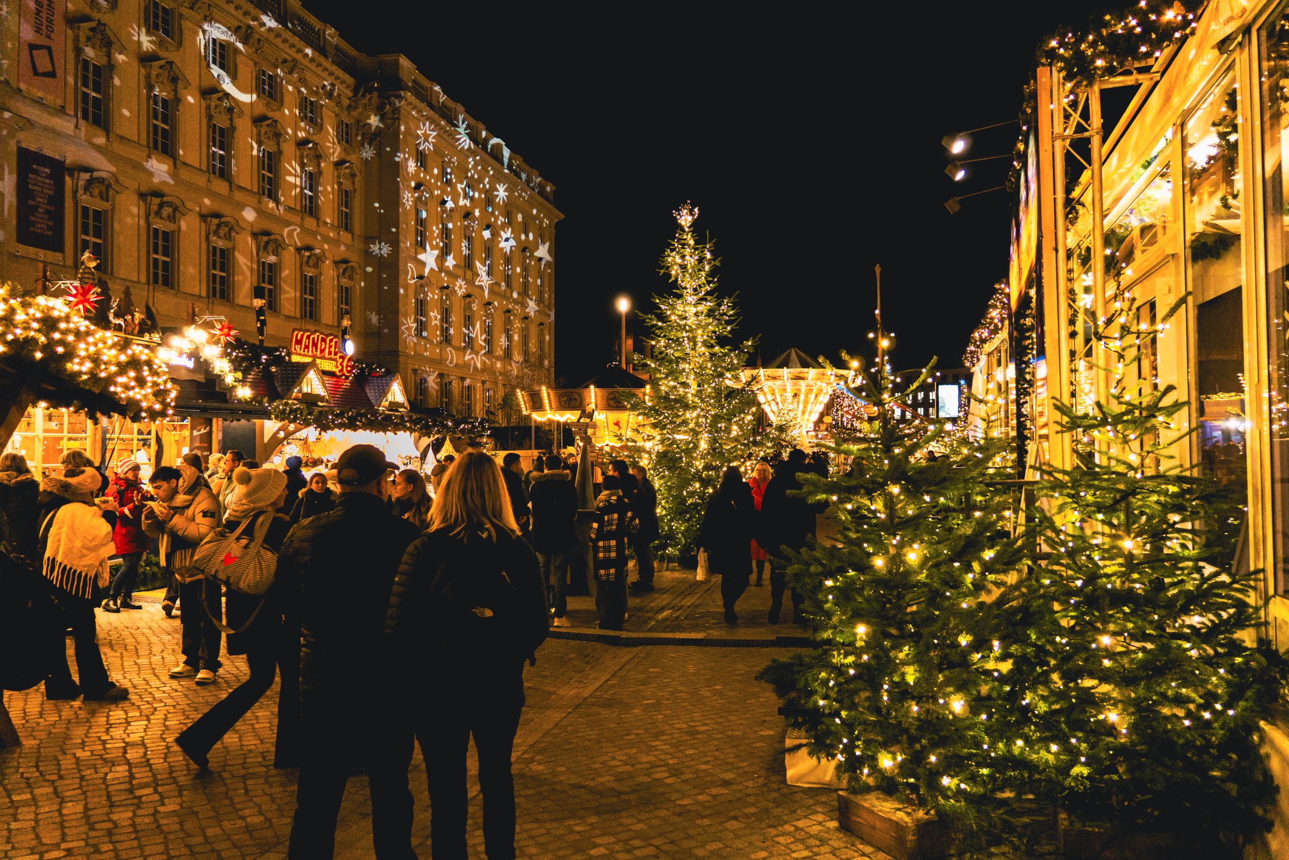 Festive night scene at a Christmas market with illuminated stalls and lively crowds.